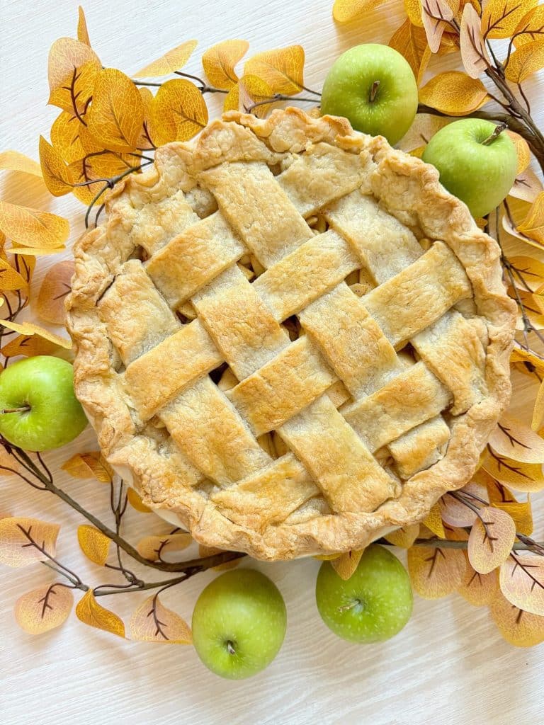 A freshly baked pie with a golden, lattice crust sits on a light wooden surface, beautifully capturing the essence of fall pies and cakes. Decorated with autumn leaves and surrounded by green apples, it exudes seasonal charm.