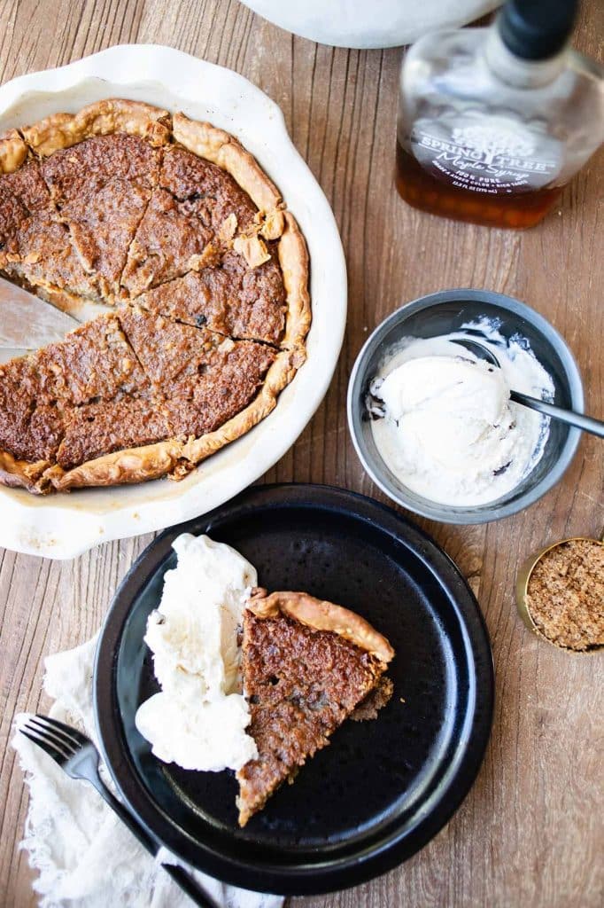 A sliced Costco Pumpkin Pie in a white dish on a wooden table, with a slice served on a black plate alongside whipped cream. A bowl of additional cream, a jar of brown sugar, and a bottle of bourbon are nearby. A fork and napkin complete this inviting copycat dessert recipe setup.