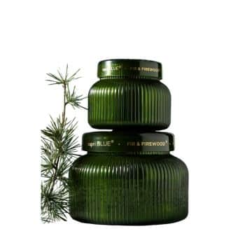 Two green candles with ribbed glass design are stacked, labeled "Capri Blue" and "Fire & Firewood." A small pine branch is placed beside them, conveying a natural and cozy theme.