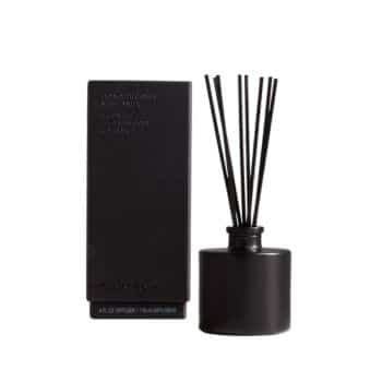 A black, cylindrical reed diffuser with long, thin sticks, next to a matching black rectangular box with text on it. The minimalist design gives a sleek and modern appearance.