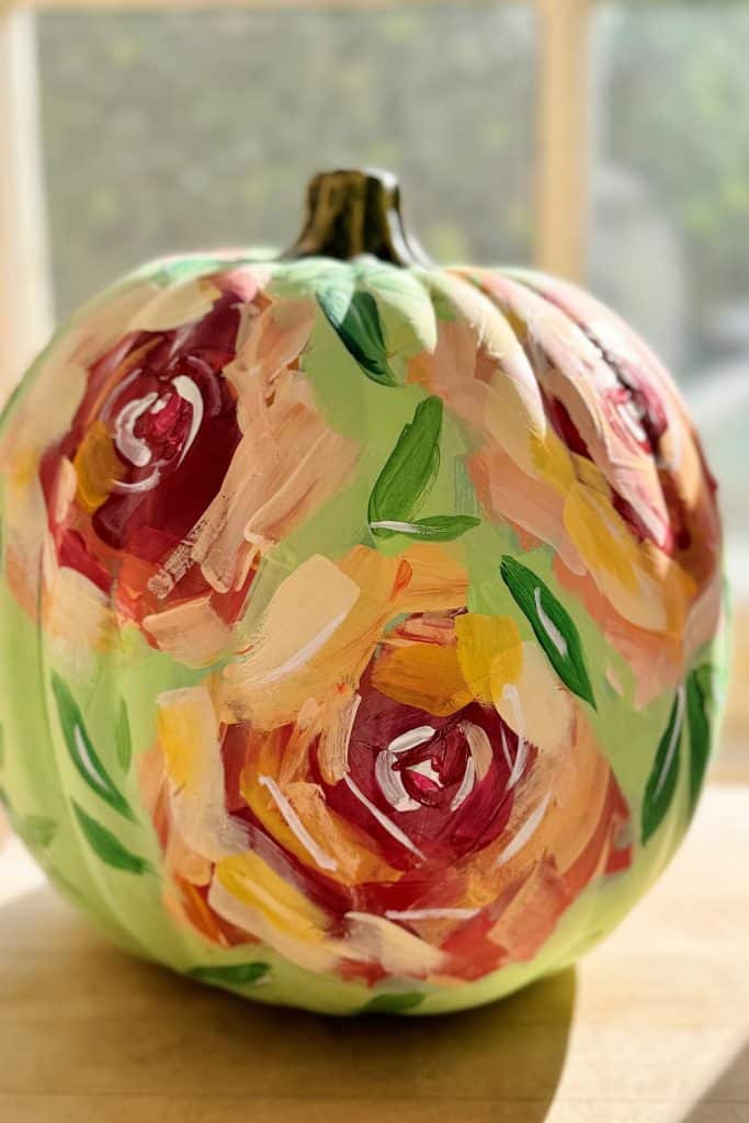 A pumpkin with flowers sits on a wooden surface, painted in vibrant floral designs of red, pink, yellow, and green. Sunlight streams in through the window behind it, highlighting this stunning DIY creation.