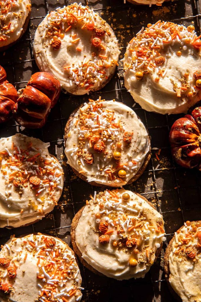 Cookies topped with white frosting and orange, yellow, and white sprinkles are arranged on a cooling rack. These delightful Fall dessert recipes are complemented by mini pumpkin-shaped pastries, adding a festive touch to the scene.