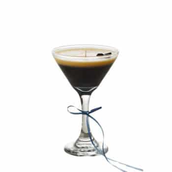 A martini glass filled with an espresso martini, displaying a rich, creamy layer on top. Three coffee beans garnish the drink, and a thin blue ribbon is tied around the stem, set against a plain white background.