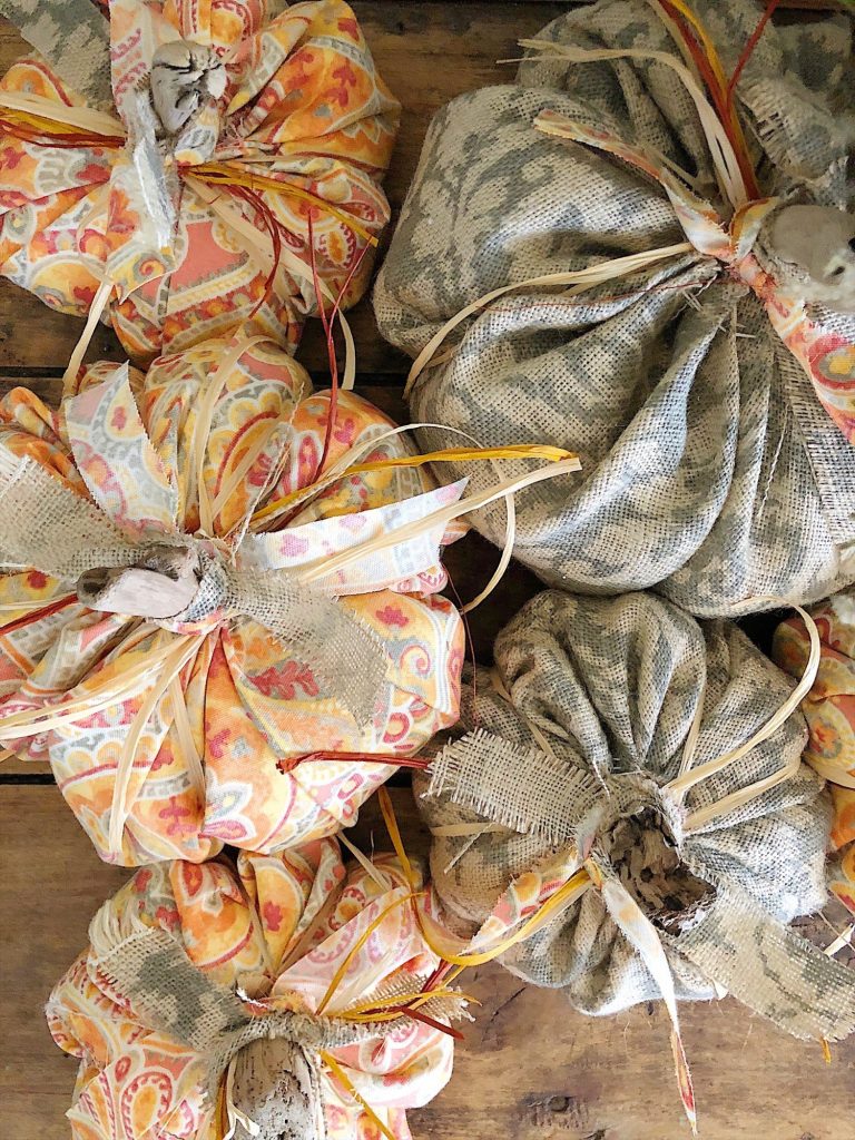 Fabric pumpkins arranged on a wooden surface exude an autumnal, rustic charm. Wrapped in vibrant orange, yellow, and beige patterned fabric with raffia bows on top, these decor pieces reflect easy pumpkin crafts perfect for bringing seasonal warmth into your space.