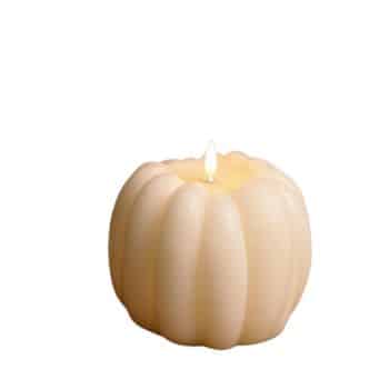 A small, cream-colored candle shaped like a pumpkin is lit, casting a soft glow. The candle has a ribbed texture and sits against a plain white background.