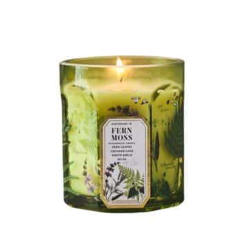 A lit candle in a green glass container with botanical designs. The label reads "Fern Moss" and "Aromatic Candle" with details on fragrance and size.