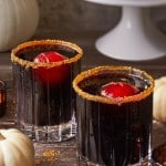 Two glass tumblers filled with a dark beverage topped with red ice spheres and rimmed with spiced sugar sit on a rustic wooden surface. Mini white pumpkins and a cake stand with a large pumpkin are in the background, creating an autumnal setting.