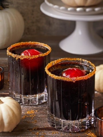 Two glass tumblers filled with a dark beverage topped with red ice spheres and rimmed with spiced sugar sit on a rustic wooden surface. Mini white pumpkins and a cake stand with a large pumpkin are in the background, creating an autumnal setting.