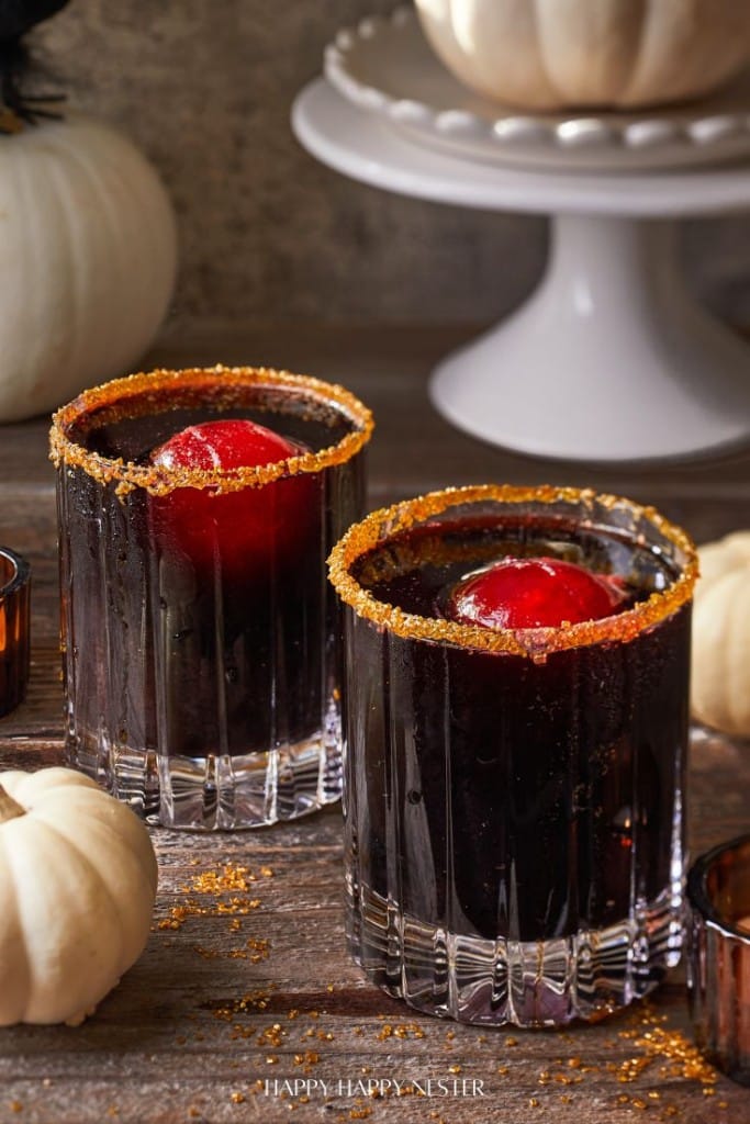 Two glass tumblers filled with a dark beverage topped with red ice spheres and rimmed with spiced sugar sit on a rustic wooden surface. Mini white pumpkins and a cake stand with a large pumpkin are in the background, creating an autumnal setting.