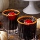 Two glass tumblers filled with a dark beverage topped with red ice spheres and rimmed with spiced sugar sit on a rustic wooden surface. Mini white pumpkins and a cake stand with a large pumpkin are in the background, creating an autumnal setting.