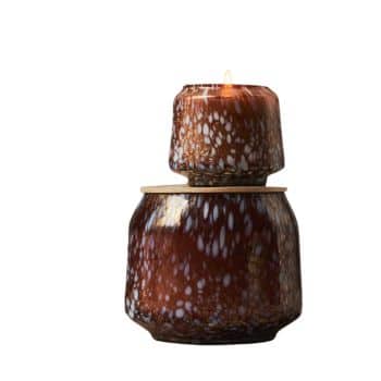 Two brown, textured glass candle holders with white speckles are stacked. The top holder has a lit candle, with a small flame casting a warm glow. The larger holder below serves as a base.