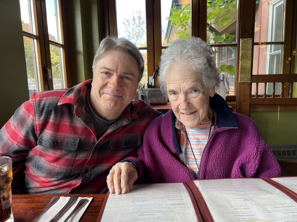 A man and an older woman sit together at a restaurant table, smiling and looking at the camera. The table has menus, a drink, and a tempting display of fall pies and cakes. The background shows large windows with a view of trees outside.