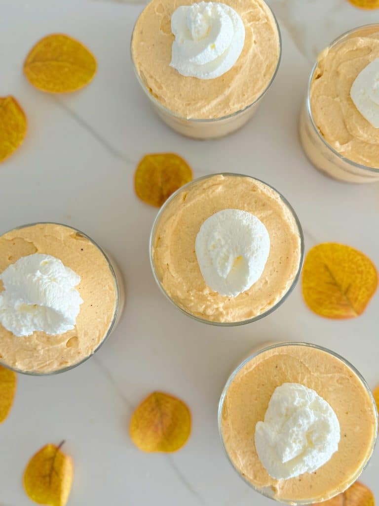 Top view of several glasses filled with creamy pumpkin mousse, a classic in fall dessert recipes, topped with dollops of whipped cream. They are arranged on a white surface, accented by scattered yellow autumn leaves for decoration.