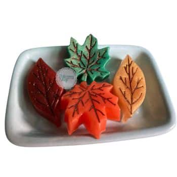 Colorful leaf-shaped soaps in red, green, orange, and yellow sit on a white rectangular dish. Each soap has a leaf vein pattern and a glittery finish.