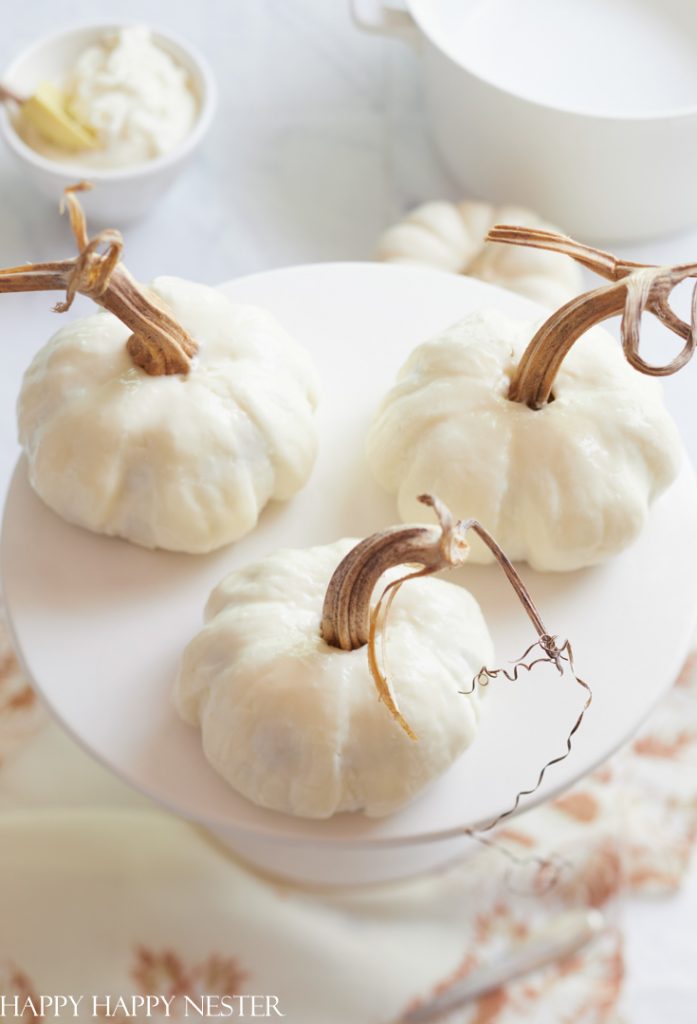 Three small white pumpkins with long stems are arranged on a white cake stand, evoking the charm of fall pies and cakes. Their rustic, curly stems add a natural touch against a soft, light-colored background with blurred elements.