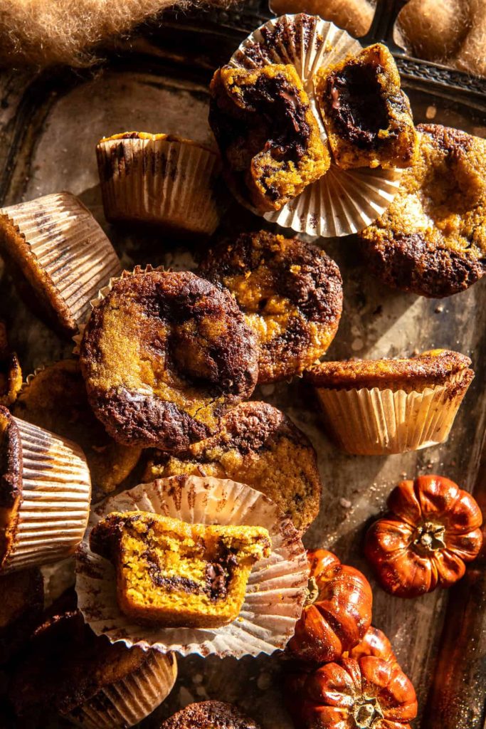 A top view of pumpkin chocolate marbled muffins on a rustic baking tray. Some muffins are broken open, revealing a moist texture inside. Mini decorative pumpkins sit beside the muffins, enhancing the fall pies and cakes theme. Shadows play across the surface.