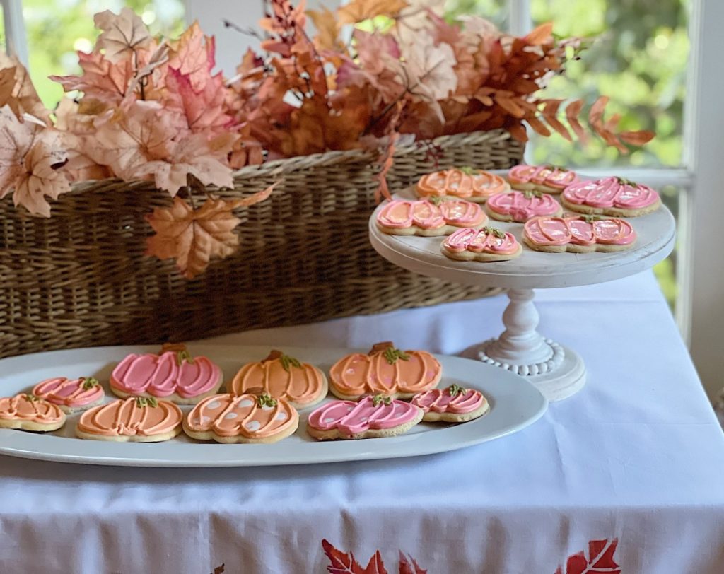 A display of pumpkin-shaped cookies in orange and pink frosting is arranged on two white platters, one elevated. Perfect Halloween treats for kids, these delights sit against a wicker basket adorned with autumn leaves on a white cloth-covered table.