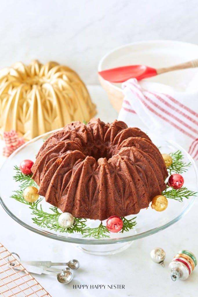 A chocolate bundt cake sits on a glass stand adorned with evergreen sprigs and colorful Christmas ornaments. A bundt pan and a red-striped towel are visible in the background.