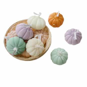 A basket filled with pastel-colored pumpkin-shaped soaps, including light green, lavender, cream, and orange. Three additional soaps are placed outside the basket. Each soap has a small tag attached.