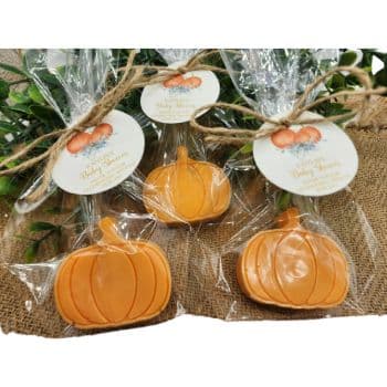 Three orange pumpkin-shaped cookies are wrapped in clear plastic and tied with twine. Each has a tag featuring pumpkins and greenery. The cookies rest on burlap with some green foliage in the background.