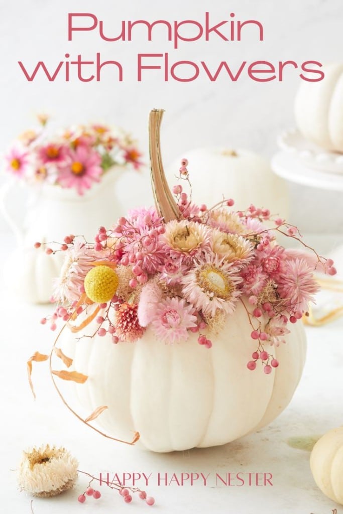 A white pumpkin is adorned with a variety of pink and yellow flowers and berries. More decorated pumpkins and a pink floral arrangement are in the background. The text reads "Pumpkin with Flowers DIY" and "Happy Happy Nester.