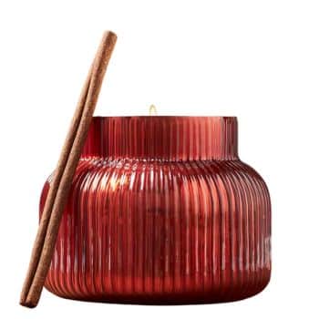 A red ribbed glass candle holder with a lit candle inside. Two cinnamon sticks lean against the side, adding a rustic touch. The glass reflects the warm glow of the candlelight.