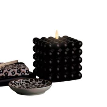 A lit black bubble cube candle sits next to two decorative plates, one with a floral pattern and the other with a leopard print design, against a white background.