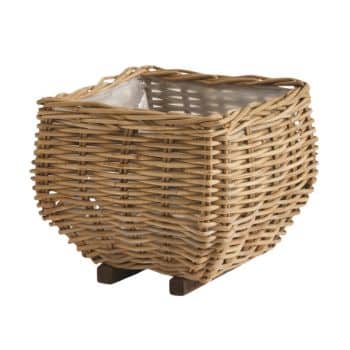 A square wicker basket with a tight weave and an open top. It features a natural, light brown color and is elevated slightly by two small wooden supports.