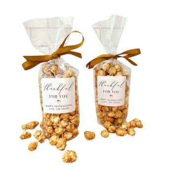 Two clear plastic bags filled with caramel popcorn are tied with brown ribbons. Each bag has a label reading "Thankful for you" and "Happy Thanksgiving, Love, The Smiths." Some popcorn pieces are scattered around the bags.