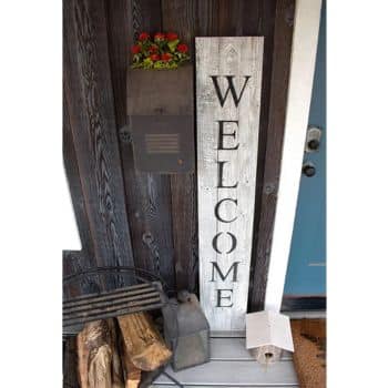 A rustic porch features a vertical "WELCOME" sign, a small lantern, chopped firewood, and a metal container. A black mailbox with colorful flowers on top is mounted on the wooden wall next to a blue door.