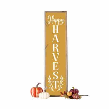 A vertical wooden sign with "Happy Harvest" written in white on an orange background. Next to the sign are small pumpkins in white and orange, and a few small decorative gourds.