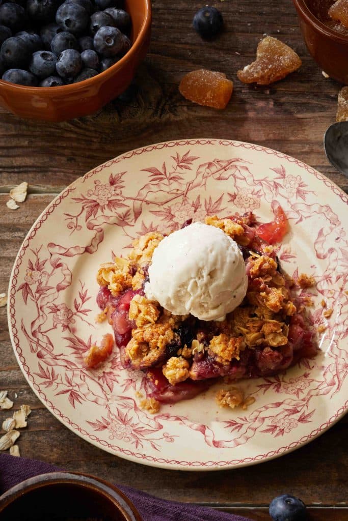 A dessert plate features a serving of blueberry crisp topped with vanilla ice cream, epitomizing fall dessert recipes. The crisp is encircled by oats and crumbs, with a bowl of fresh blueberries and ginger pieces nearby, all set on a rustic wooden table.