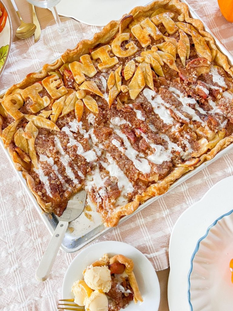 A rectangular pie reminiscent of a Costco classic, with a crumbly topping and drizzled icing, sits on a tray. The crust boasts decorative leaves and the words "Pie Night." A slice is served with ice cream atop a plaid tablecloth, capturing the essence of a beloved pumpkin pie recipe.