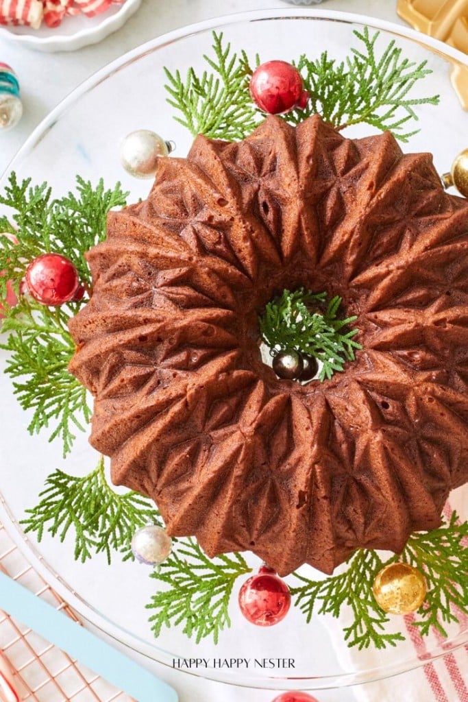 A decorative bundt cake, inspired by banana bread recipes using oil, sits on a glass stand, encircled by evergreen branches and festive red, gold, and silver ornaments. The cake features a striking geometric pattern and is beautifully viewed from above.