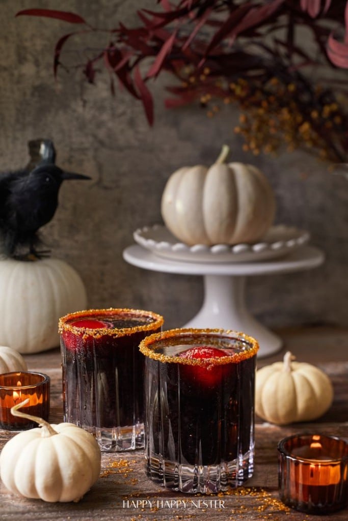 Two black cocktail drinks in sugar-rimmed glasses are surrounded by small white pumpkins and amber tea lights. A white pumpkin sits on a cake stand, with a decorative black crow perched nearby on another pumpkin. Autumnal leaves add to the festive setting.