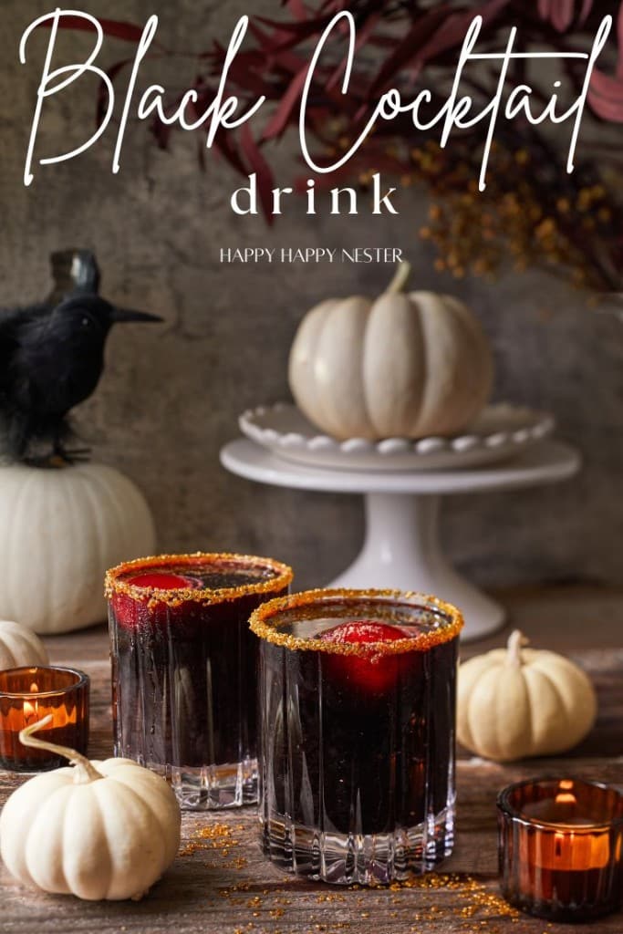 A Halloween-themed setting features two black cocktail drinks with dark allure, garnished with orange sugar rims. They are surrounded by small white pumpkins and lit candles, while a black crow figurine perches atop a larger pumpkin in the background, adding an eerie charm to the scene.