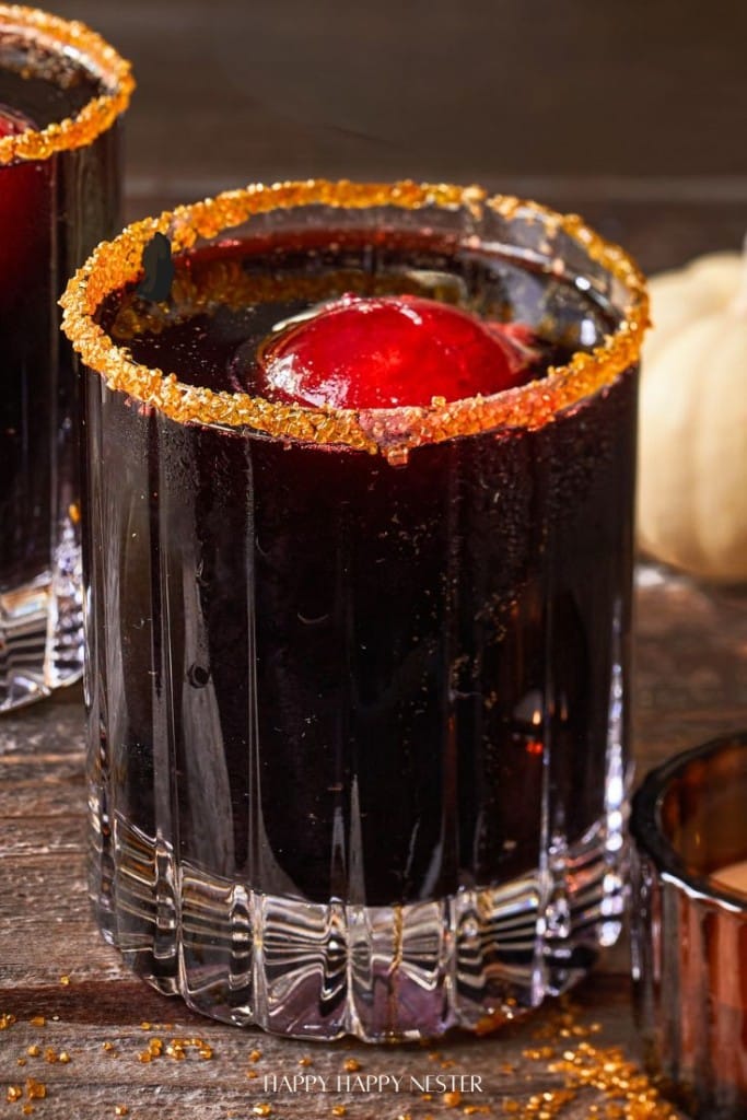 A close-up of a dark, black cocktail drink in a glass with a sugared rim and a cherry garnish. The drink is set on a wooden surface, accented by the soft contrast of a white pumpkin in the background.