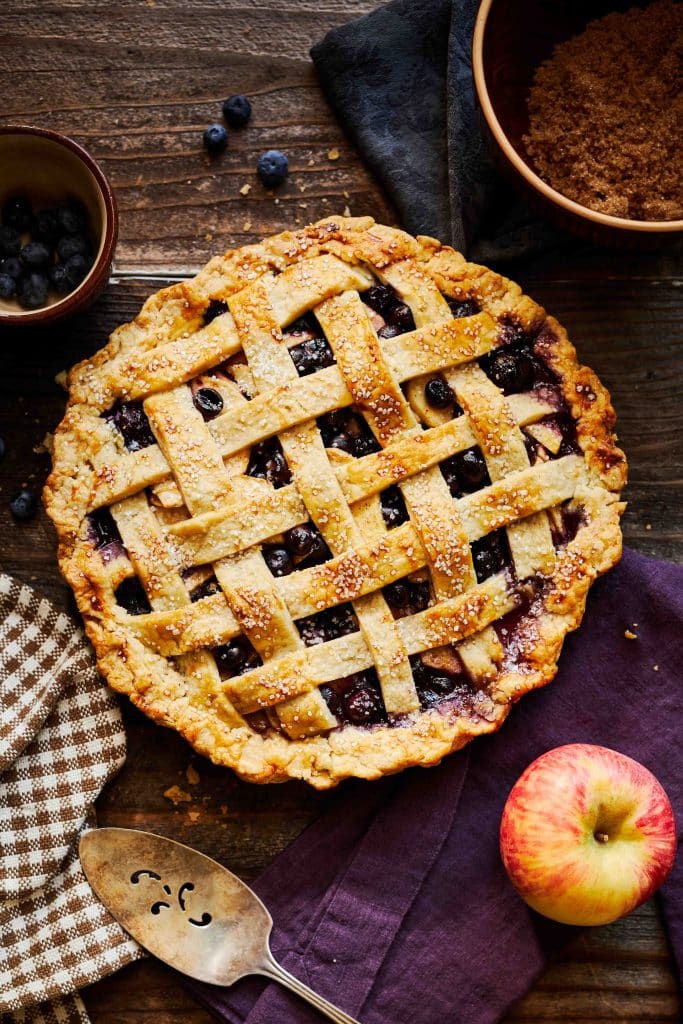 A freshly baked blueberry pie with a golden lattice crust sits on a wooden table. Nearby, blueberries and brown sugar hint at the delicious blend typical of fall pies and cakes. A checkered cloth, decorative pie server, and an apple on a purple napkin complete the cozy scene.