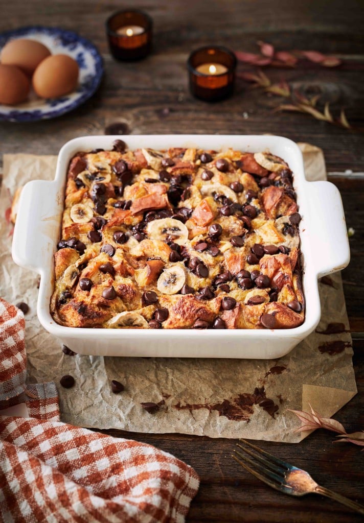 Square white baking dish filled with baked banana and chocolate chip bread pudding on parchment paper—a perfect addition to your fall dessert recipes. Whole eggs in the background, candles lit on a rustic wooden table, and a checkered cloth nearby complete the cozy scene.