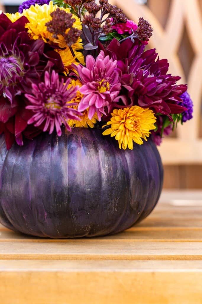 A decorative DIY arrangement showcases deep purple and yellow flowers set inside a dark purple pumpkin vase, placed on a wooden surface. The background reveals a soft-focus pattern of carved wood, enhancing the creative charm of this pumpkin with flowers display.