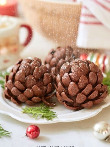 Three chocolate pinecone-shaped treats dusted with powdered sugar are displayed on a white plate. Festive decorations, including greenery and ornaments, surround the plate.