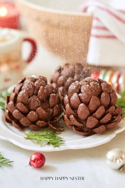 Three chocolate pinecone-shaped treats dusted with powdered sugar are displayed on a white plate. Festive decorations, including greenery and ornaments, surround the plate.