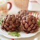 Three chocolate pinecone-shaped treats dusted with powdered sugar are displayed on a white plate. Festive decorations, including greenery and ornaments, surround the plate.