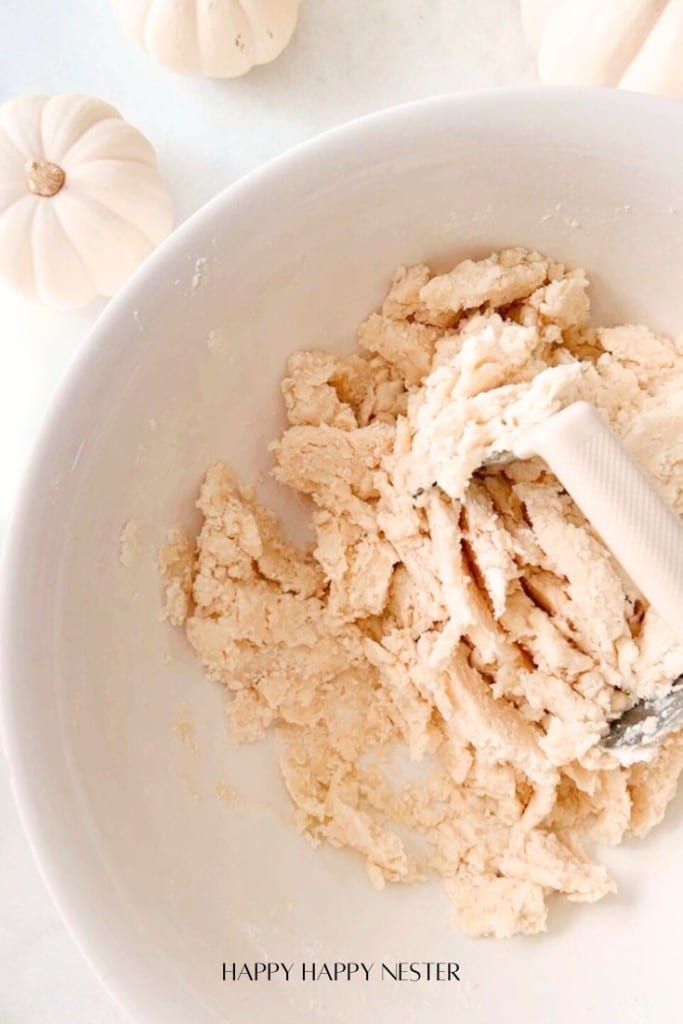 A bowl filled with crumbly pastry dough, with a spatula resting on the side. The background features small white pumpkins. The image has a soft, light aesthetic. Text at the bottom reads "HAPPY HAPPY NESTER.