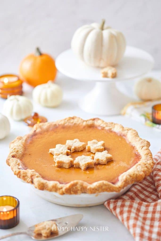 A pumpkin pie topped with star-shaped pastry sits on a table, embodying the essence of fall pies and cakes. In the background, white and orange pumpkins grace a cake stand, surrounded by cozy candles and a checkered cloth, enhancing the warm autumn atmosphere.