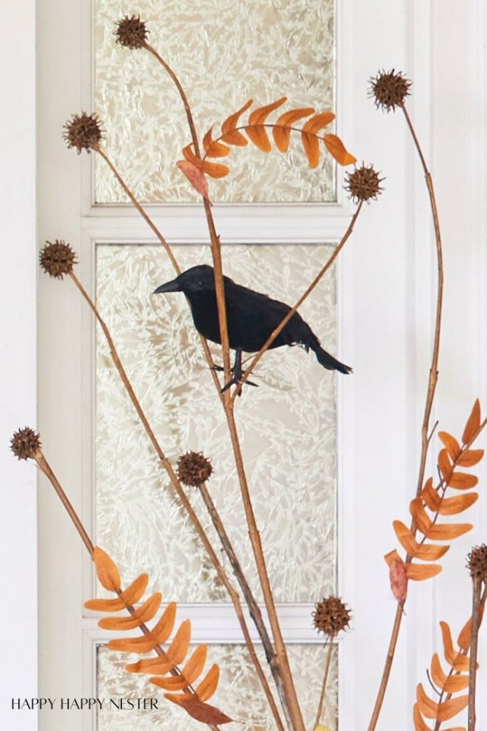 A black bird is perched on a decorative branch with orange, curved leaves and round seed pods. The background features a frosted glass door with a textured design.