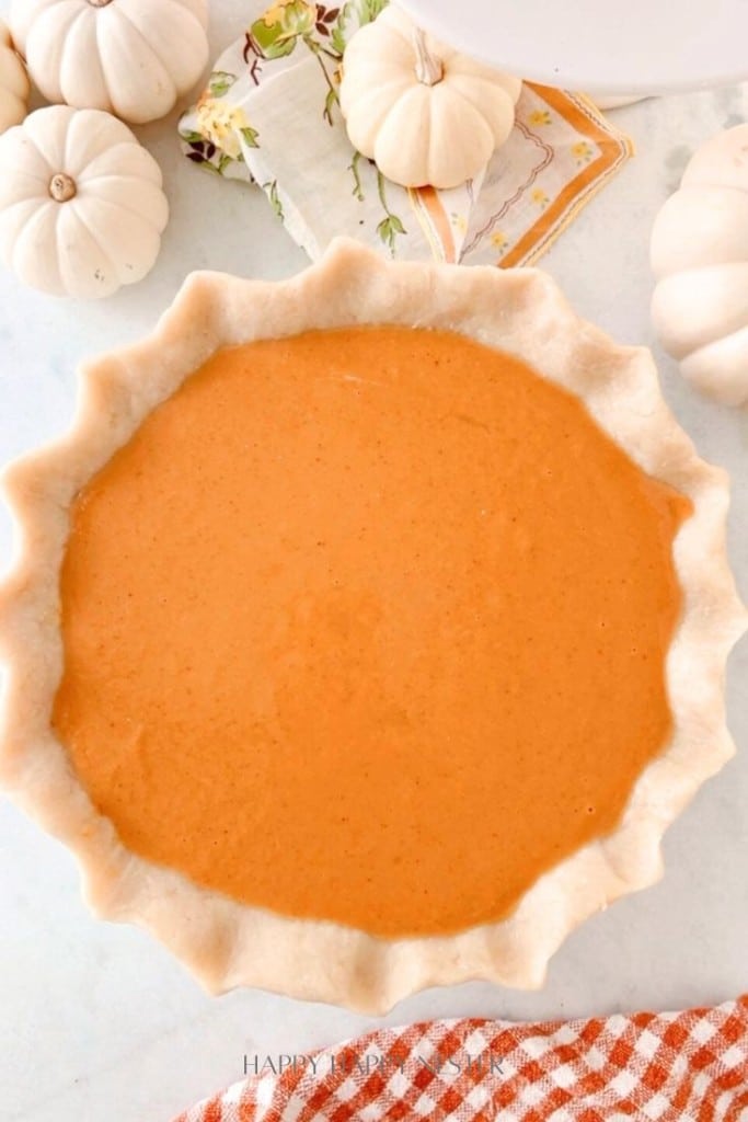 A pumpkin pie filling, reminiscent of a Costco Pumpkin Pie Recipe copycat, rests in an unbaked crust on a white surface. It's surrounded by small white pumpkins and decorative napkins, with a checkered cloth in the bottom right corner adding cozy charm.