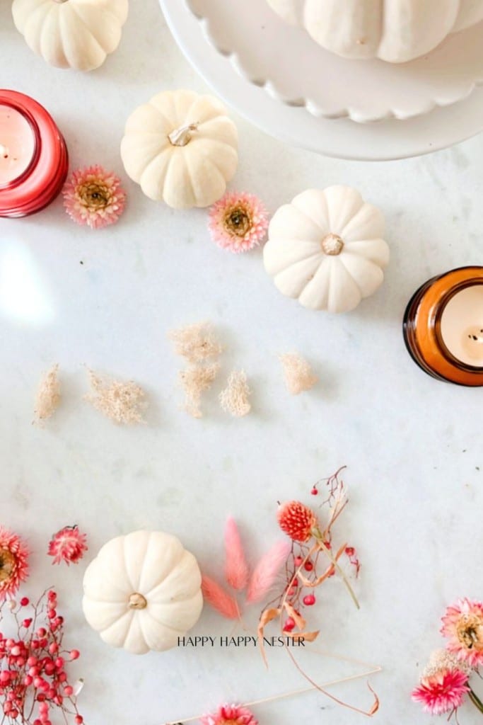 A light and airy autumn-themed flat lay with small white pumpkins, pink and red dried flowers, and lit candles on a white surface. The text "Happy Happy Nester" is subtly displayed at the bottom.