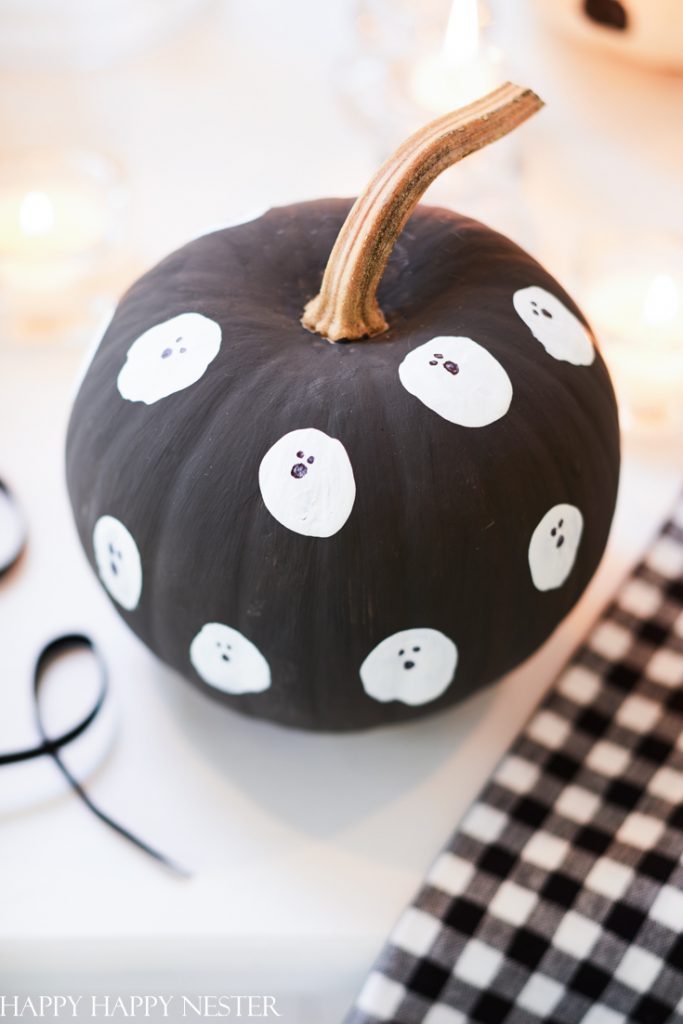 A black pumpkin, an easy pumpkin craft, is painted with small white ghost faces and features a brown stem. It rests surrounded by candles atop a black-and-white checkered cloth.