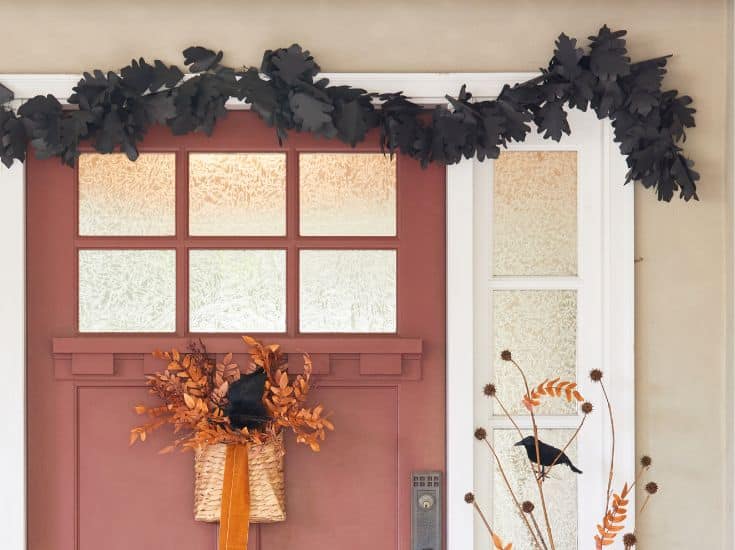 A red door decorated for autumn with a basket of orange leaves and a black crow. Black leaf garlands hang above the door, and additional autumn foliage and a black crow are arranged to the side.